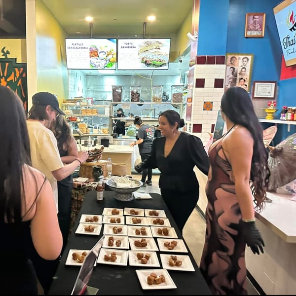 People sampling small dishes of Thai food at a restaurant event with an open kitchen.