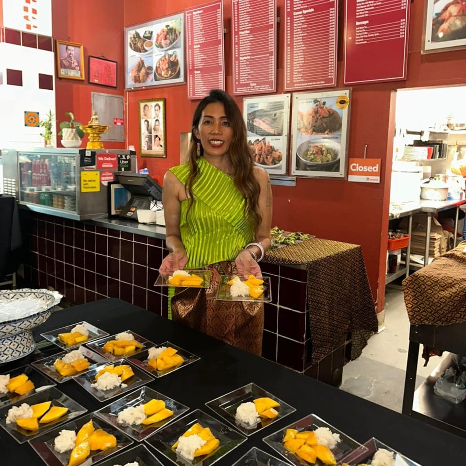 A woman in traditional Thai attire holds two plates of mango sticky rice in a restaurant.